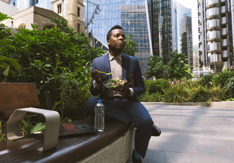 Älterer Geschäftsmann sitzt mit einer Schüssel Salat auf einer Bank im Büropark - OIPF03824