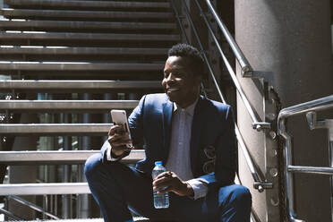 Lächelnder Geschäftsmann mit Wasserflasche und Smartphone auf Stufen sitzend - OIPF03828