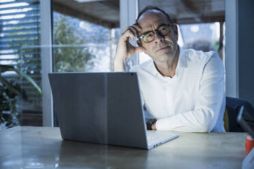 Thoughtful mature freelancer with head in hand near laptop at home office - UUF31058