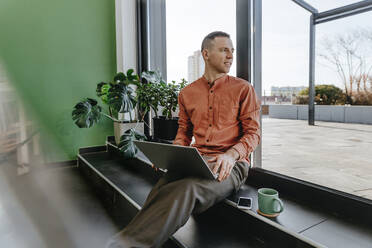 Freelancer sitting with laptop near glass door - YTF01630