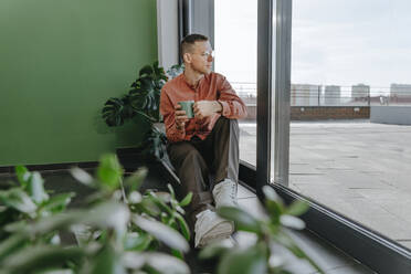 Man sitting with coffee cup near glass door - YTF01631