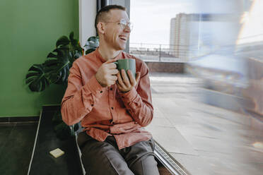 Happy man sitting with coffee cup near glass door - YTF01632