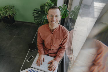 Lächelnder Freiberufler mit Laptop, der in der Nähe einer Glastür sitzt - YTF01634