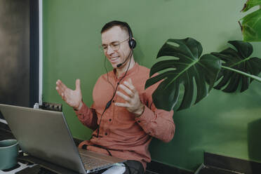 Smiling freelancer using headset and laptop for video call in front of green wall - YTF01639