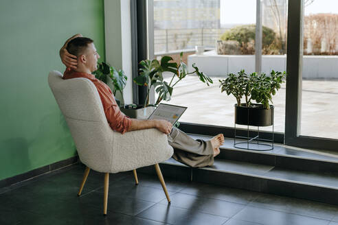 Nachdenklicher Freiberufler auf einem Sessel sitzend mit Laptop im Home Office - YTF01650