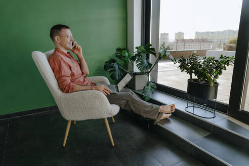 Lächelnder Freiberufler, der auf einem Sessel sitzt und über sein Smartphone spricht - YTF01651