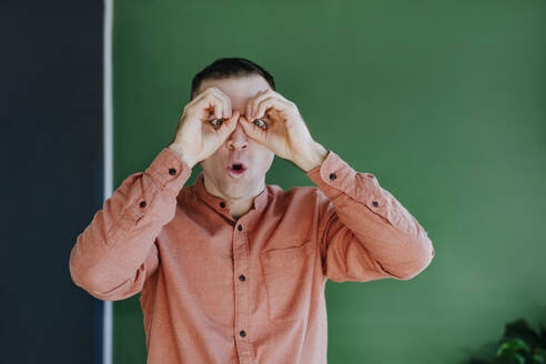 Surprised man staring through finger binoculars in front of green wall - YTF01665