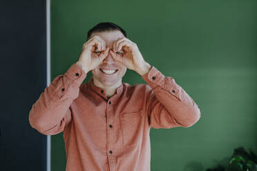 Playful man looking through finger binoculars in front of green wall - YTF01666