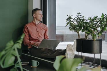 Thoughtful freelancer sitting with laptop near glass door at home office - YTF01673