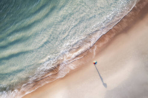 Luftaufnahme einer Person, die an einem weißen Sandstrand steht und einen bunten Regenschirm hält, mit klarem, blauem Wasser und Langzeitbelichtungseffekt, Port Noarlunga, South Australia, Australien. - AAEF25238