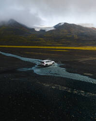 Drohnenaufnahme eines weißen Autos, das durch einen kleinen Fluss vor einem Berg im Hochland von Island fährt. - AAEF25926