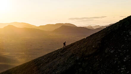 Aerial drone view of a silhouette person walking uphill on Blahylur Crater during sunset in the highlands of Iceland. - AAEF25928