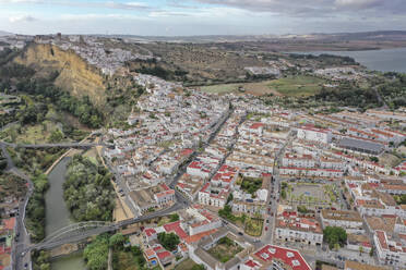 Luftaufnahme der Stadt Arcos de la Frontera mit einem See im Hintergrund und ihren weißen Häusern und roten Dächern, die sich an senkrechten Klippen in Cádiz, Andalusien, Spanien, emporheben. - AAEF25940