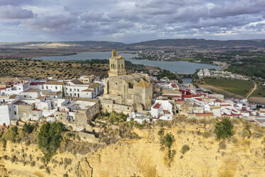 Luftaufnahme einer Kathedrale, Iglesia de San Pedro, mit einem See im Hintergrund, an einem bewölkten Tag auf einem Sandsteinrücken in der Stadt Arcos de la Frontera in Cadiz, Andalusien, Spanien. - AAEF25942
