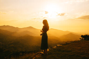 Eine einsame Frau steht mit Blick auf eine Bergkette im warmen Licht der untergehenden Sonne - ADSF52610