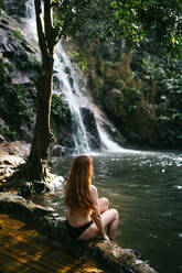 A person is sitting serenely by a secluded waterfall surrounded by lush greenery - ADSF52611