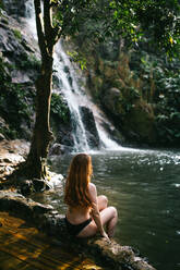 Eine Person sitzt in aller Ruhe an einem abgelegenen Wasserfall, umgeben von üppigem Grün - ADSF52611