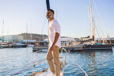 Lächelnder bärtiger Mann in weißem Hemd, der von einer Yacht vor blauem Himmel und Meer wegschaut - ADSF52676