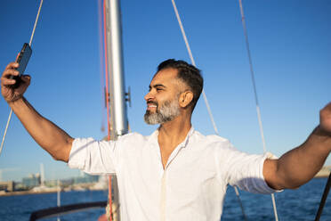 Lächelnder bärtiger Mann in weißem Hemd, der ein Selfie mit seinem Handy macht, während er auf einer Yacht vor blauem Meer und Himmel steht - ADSF52677