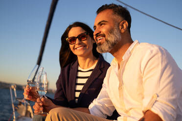 Smiling couple sitting on yacht and clinking wineglasses while enjoying romantic time against in waving ocean - ADSF52679