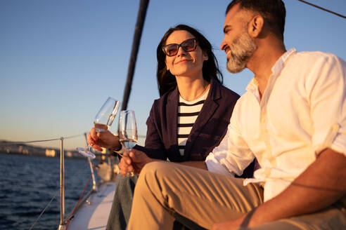 Lächelndes Paar sitzt auf einer Yacht und stößt mit Weingläsern an, während es die romantische Zeit im wogenden Meer genießt - ADSF52680