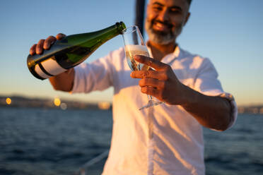 Lächelnder Mann in weißem Hemd auf einer Yacht stehend und Wein in ein Weinglas einschenkend, während er seine Zeit mit verschwommenem Meerwasser verbringt - ADSF52681