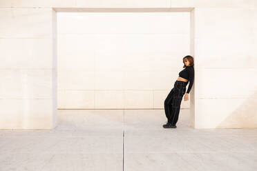 A stylish young woman in fashionable black attire confidently poses against a stark, minimalist architectural structure, reflecting a contemporary urban vibe - ADSF52683