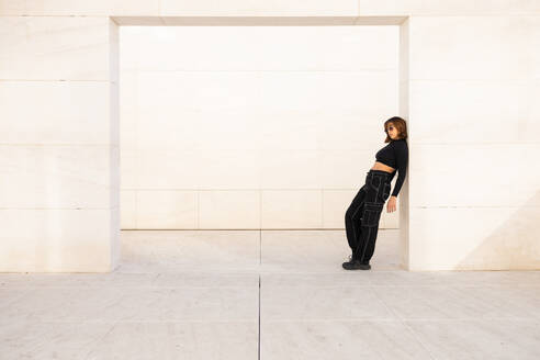 Eine stilvolle junge Frau in modischer schwarzer Kleidung posiert selbstbewusst vor einer strengen, minimalistischen architektonischen Struktur, die eine zeitgenössische urbane Atmosphäre widerspiegelt - ADSF52683
