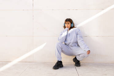 A young woman in a comfortable blue tracksuit and black sneakers sits against a minimalist background, listening to music with blue headphones - ADSF52685