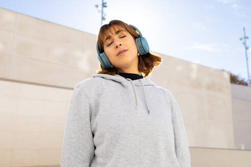 Eine ruhige junge Frau mit grauem Kapuzenpulli und blauen Kopfhörern schließt zufrieden die Augen und genießt die Musik in einer städtischen Umgebung - ADSF52686