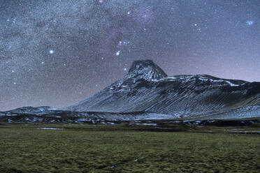 Sternenübersäter Nachthimmel mit der Milchstraße über einem schneebedeckten Berg und einem grasbewachsenen Feld im isländischen Winter - ADSF52704