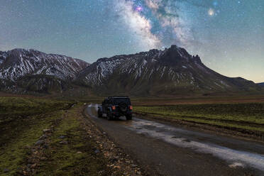 Auf einem Schotterweg geparkter Geländewagen vor der Kulisse schneebedeckter Berge unter der Milchstraße in Island bei Nacht - ADSF52705
