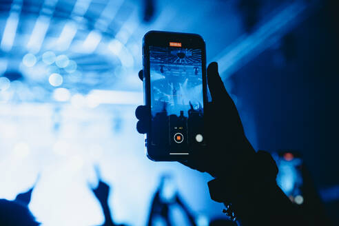 Eine silhouettierte Hand hält ein Smartphone hoch und nimmt eine Live-Konzertszene auf, die durch den Bildschirm vor dem Hintergrund der Bühnenbeleuchtung und des jubelnden Publikums zu sehen ist - ADSF52711