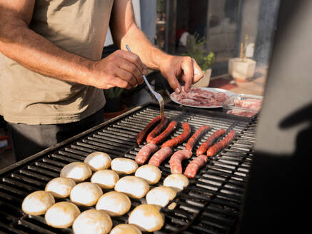 Von oben von einem anonymen erwachsenen männlichen Koch, der in der Nähe eines Grillrosts mit Würstchen im Tageslicht steht und eine Stahlgabel zum Wenden benutzt, während er in einer offenen Küche arbeitet - ADSF52714