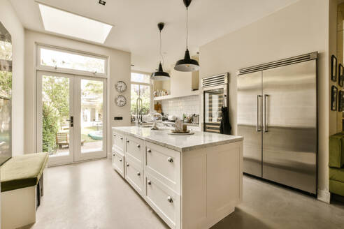 Eine helle und geräumige moderne Küche mit weißen Schränken, Marmorarbeitsplatten und Edelstahlgeräten. Viel Sonnenlicht erfüllt den Raum durch große Fenster, die den Blick auf den grünen Garten freigeben. - ADSF52725