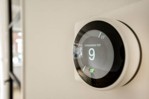 Nahaufnahme eines digitalen, intelligenten Thermostats, der an einer Wand montiert ist und eine Temperatureinstellung mit einem Weichzeichner-Hintergrund zeigt. - ADSF52735