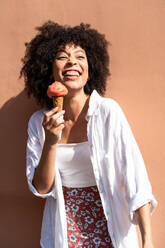 Curly haired black woman in white enjoys a strawberry ice cream cone against a warm peach wall looking away - ADSF52746