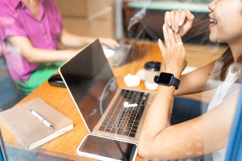 Abgeschnittene, nicht erkennbare, lächelnde Frau, die eine Smartwatch trägt und sich mit einem Kollegen in einem Café unterhält, während sie an ihrem Laptop arbeitet, Fernarbeit in Chiang Mai, Thailand - ADSF52747