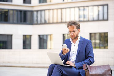 Businessman with laptop using smart phone in the city - PUF02045