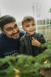 Smiling man looking at Christmas tree with son at home - ANAF02664