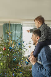 Cheerful man carrying son on shoulders near Christmas tree at home - ANAF02670