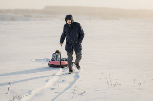 Mann zieht Junge auf aufblasbarem Schlauch im Schnee sitzend - ANAF02683