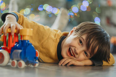 Glücklicher Junge liegt auf dem Boden und spielt mit einem Zugspielzeug zu Hause - ANAF02694