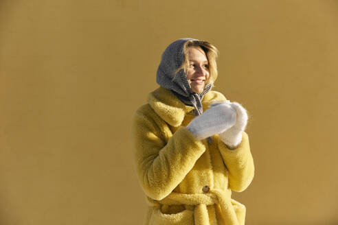 Glückliche junge Frau in Pelzmantel und mit Kaffeetasse vor gelber Wand - SANF00161