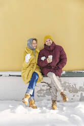 Lächelndes Paar hält Kaffeetasse und sitzt in der Nähe von gelben Wand im Winter - SANF00167