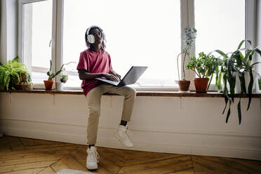 Nachdenklicher Mann sitzt zu Hause auf der Fensterbank mit Laptop - NLAF00215