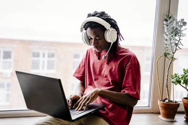 Man wearing wireless headphones and using laptop near window at home - NLAF00217