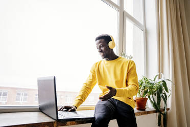 Mann mit drahtlosen Kopfhörern und Videogespräch am Laptop in der Nähe des Fensters - NLAF00219