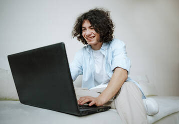 Glücklicher junger Mann mit lockigem Haar, der auf dem Sofa zu Hause einen Laptop benutzt - NLAF00227