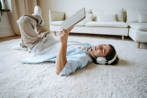 Junger Mann auf Teppich liegend und mit Tablet-PC zu Hause - NLAF00231
